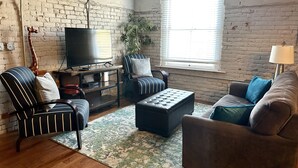 Ein Wohnzimmer mit Backsteinwand, Fernseher, Sofa, Sessel und einer Lampe.