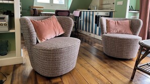 Uma sala de estar acolhedora com duas poltronas estampadas, piso de madeira, cozinha com armários listrados e uma janela com persianas.