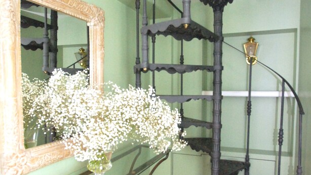 Uma escadaria em espiral com grades de metal elaboradas, um espelho com moldura ornamentada e um vaso com flores brancas sobre um aparador.
