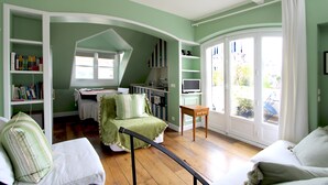Quarto com mesa de jantar, cadeiras, sofá, mesa pequena e televisão. Há uma janela com cortinas e vista para o exterior.