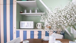Uma área de refeições com mesa de madeira posta para dois, um prato branco com macarons e uma xícara branca. Há um micro-ondas branco em uma prateleira acima da mesa, e um vaso com flores brancas à direita.