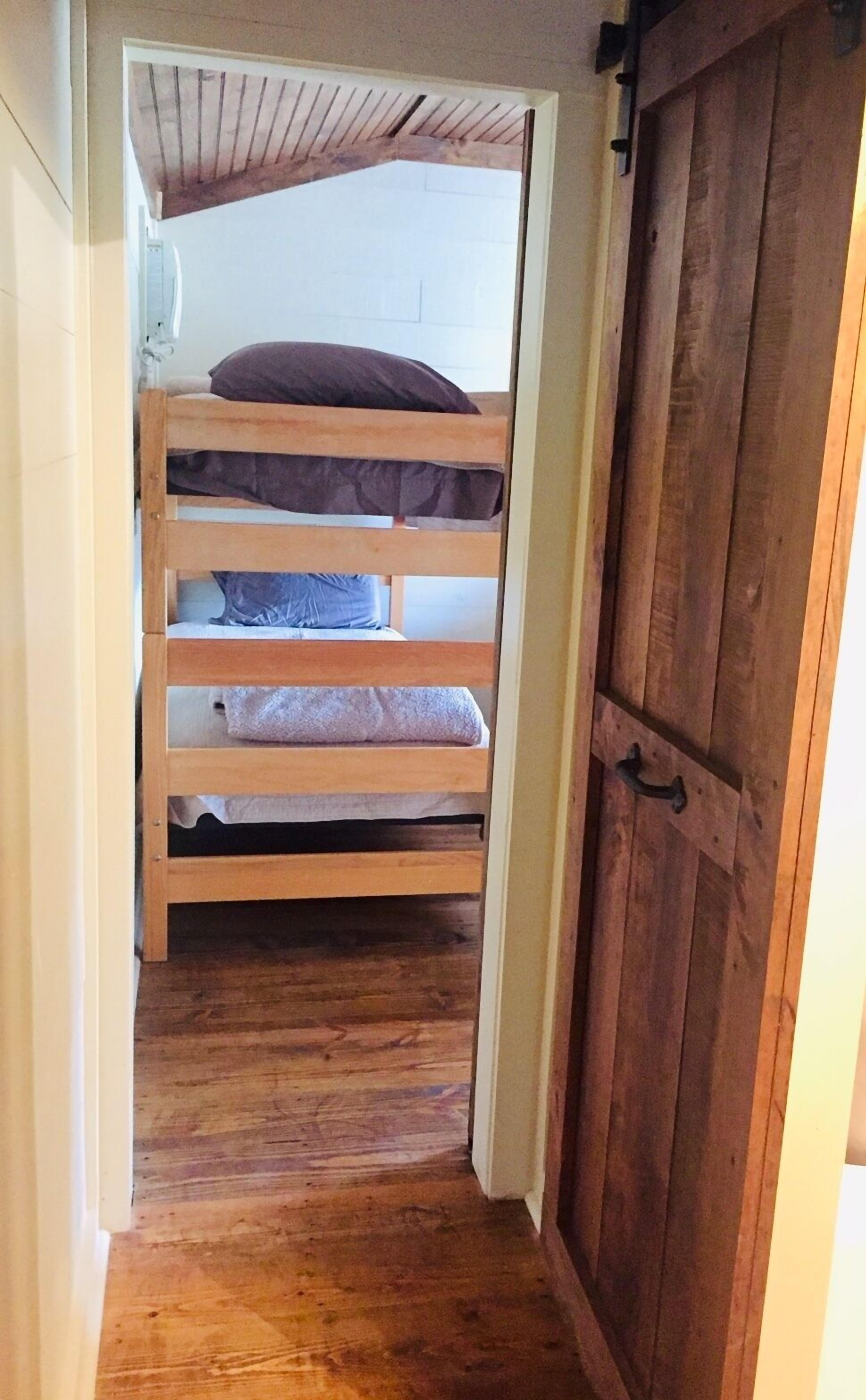 Un couloir étroit mène à une pièce où se trouve un lit superposé, et une porte en bois se trouve sur le côté droit.