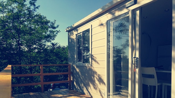 Une terrasse en bois avec une rambarde, une porte vitrée donnant sur un espace intérieur où se trouve une chaise blanche, et une vue sur des arbres en arrière-plan.