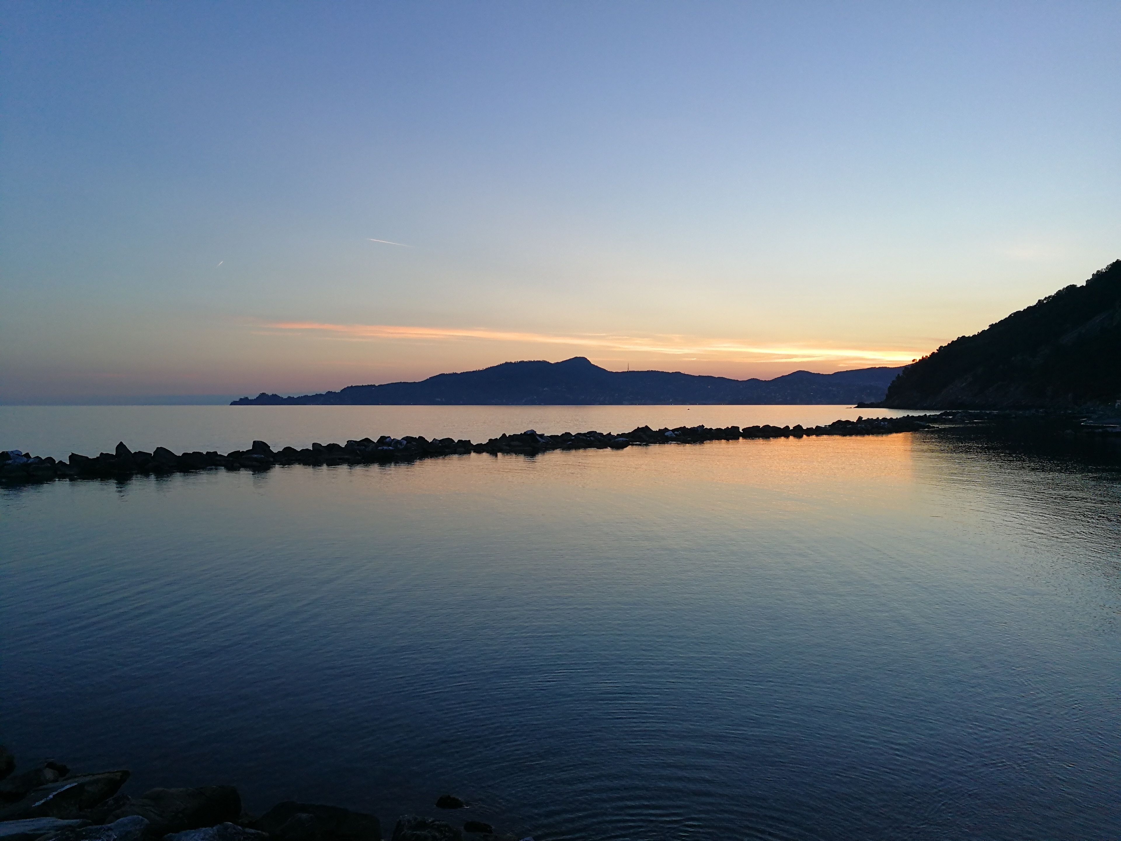 A calm body of water with a rocky breakwater and distant mountains during sunset.