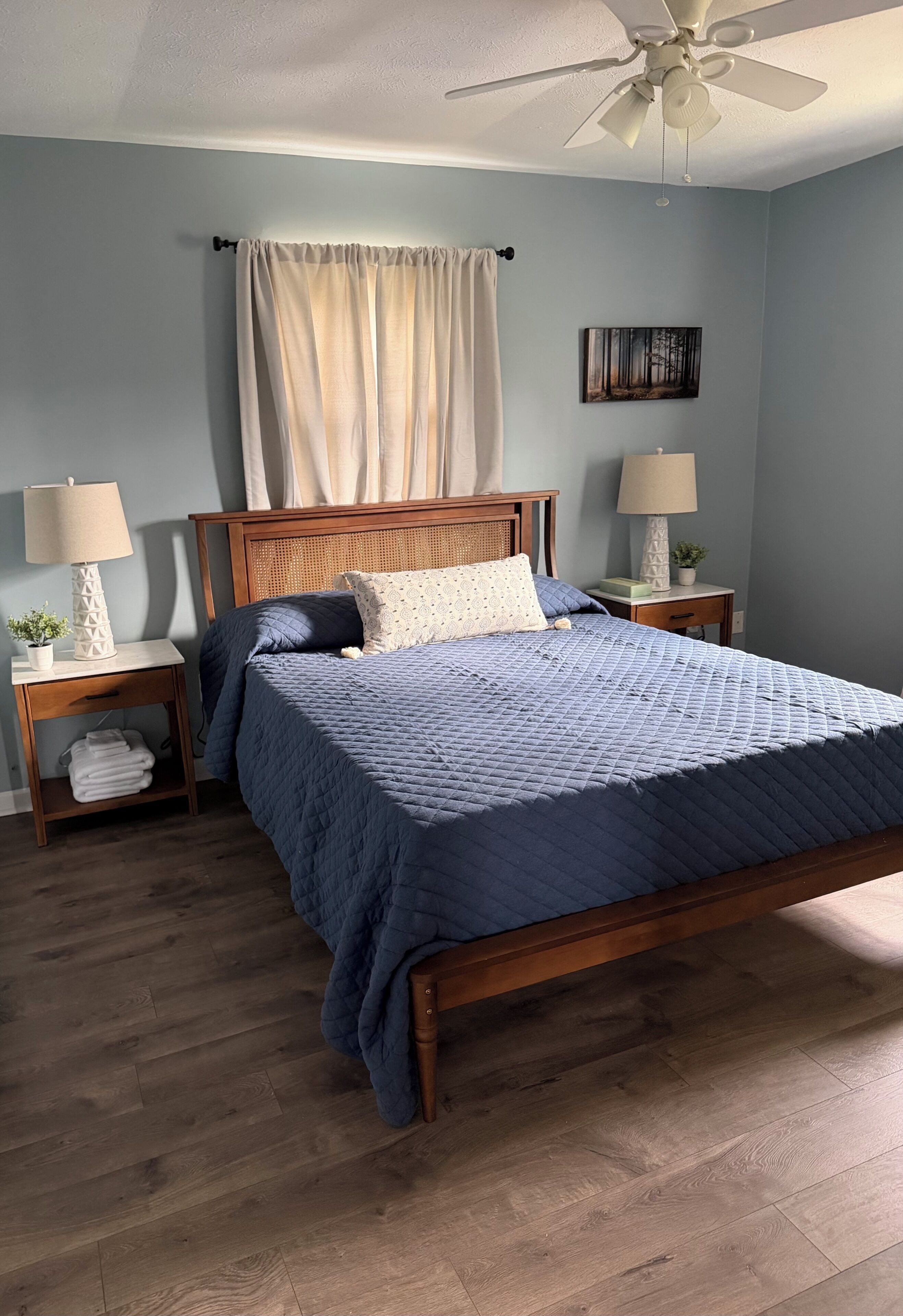 Une chambre à coucher avec un lit en bois, deux tables de chevet avec des lampes, un ventilateur de plafond et un tableau accroché au mur.