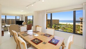 A spacious living room with a large table, comfortable seating, a flat-screen TV, and a view of the beach and cityscape through large windows. - Gulf & Bay Club, TopFloor End Unit 1947sqft incl Lanai, (Siesta Key)