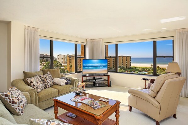 A modern living room with a large TV, comfortable seating, and a view of the beach.