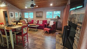 A cozy cabin interior with a dining area, a living room with a fireplace, wooden floors, and a stone wall.