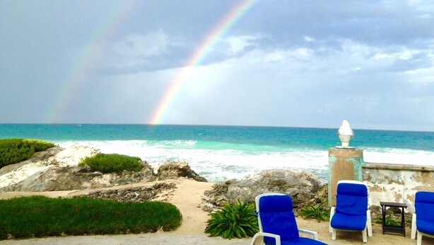 A beach view with a rainbow, blue lounge chairs, and a stone pillar. - Casa Rosa -Beautiful Ocean Front Home - Studio Apartment (Isla Mujeres)