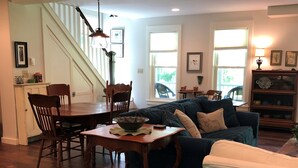 A spacious living room with a dark blue sofa, a dining table with chairs, a wooden coffee table, and a staircase leading to an upper level. - Lovely Cottage That Is Completely New With All The Luxuries You Would Want!   (Great Valley)