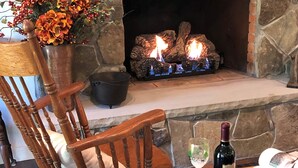 A stone fireplace with a fire, a wooden chair, a vase with flowers, a wine glass, and a bottle. - Lovely Cottage That Is Completely New With All The Luxuries You Would Want!   (Great Valley)