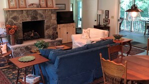 A cozy living room with a stone fireplace, a blue sofa, wooden furniture, and a view of the outdoors. - Lovely Cottage That Is Completely New With All The Luxuries You Would Want!   (Great Valley)