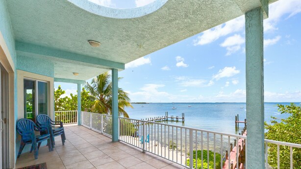 Ein überdachter Balkon mit Meerblick, ausgestattet mit Geländer, Stühlen und einer Tür zu einem Innenraum.