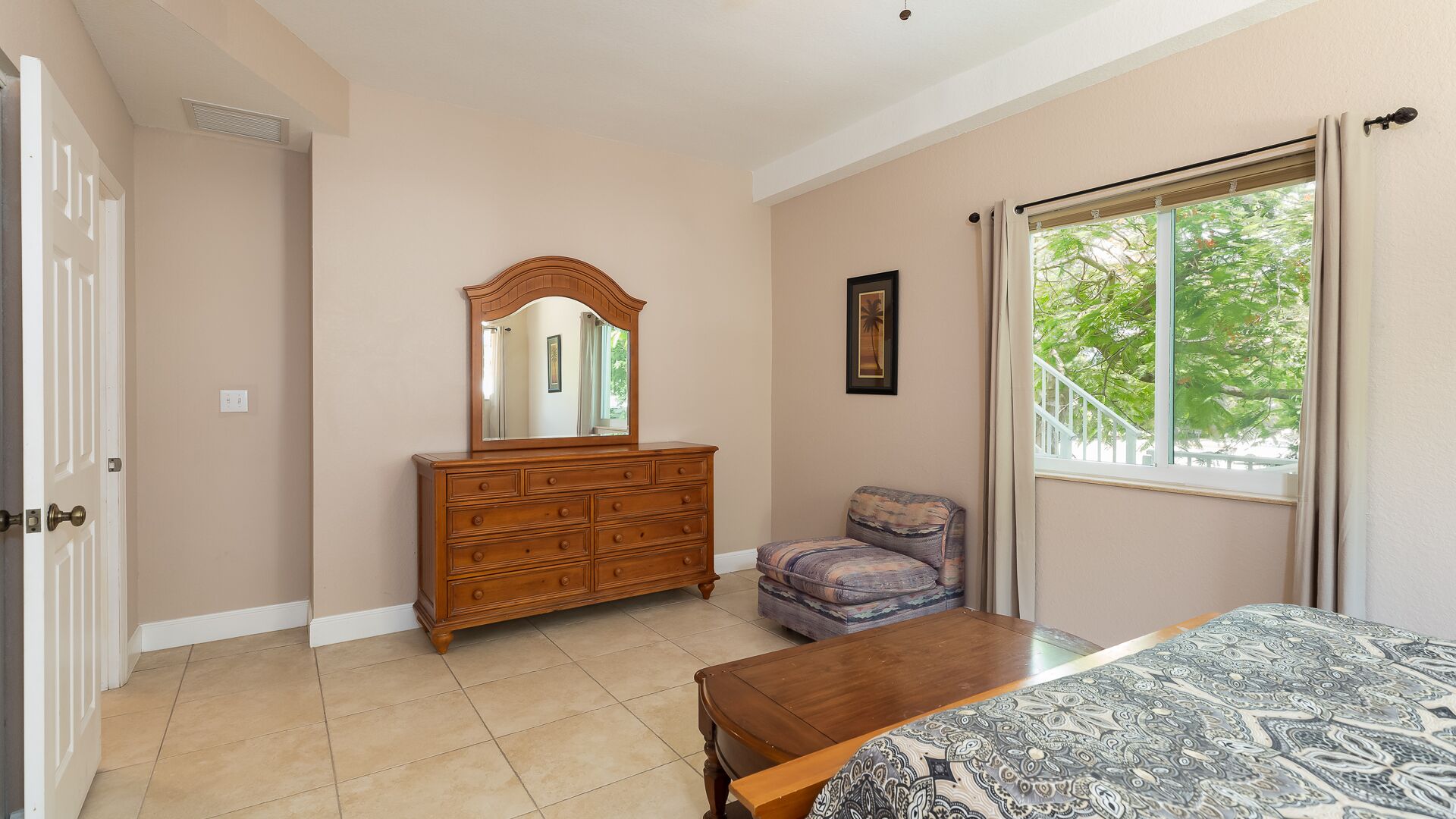 A bedroom with a wooden dresser, a chair, a bed with a patterned bedspread, a window with a view of trees, and a door.