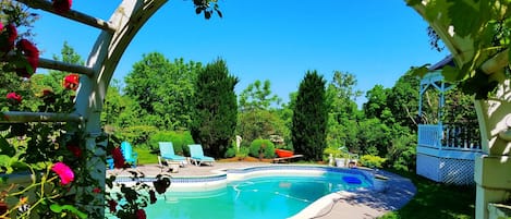 A swimming pool with a white pergola and red flowers in the foreground, surrounded by green trees and a clear blue sky.