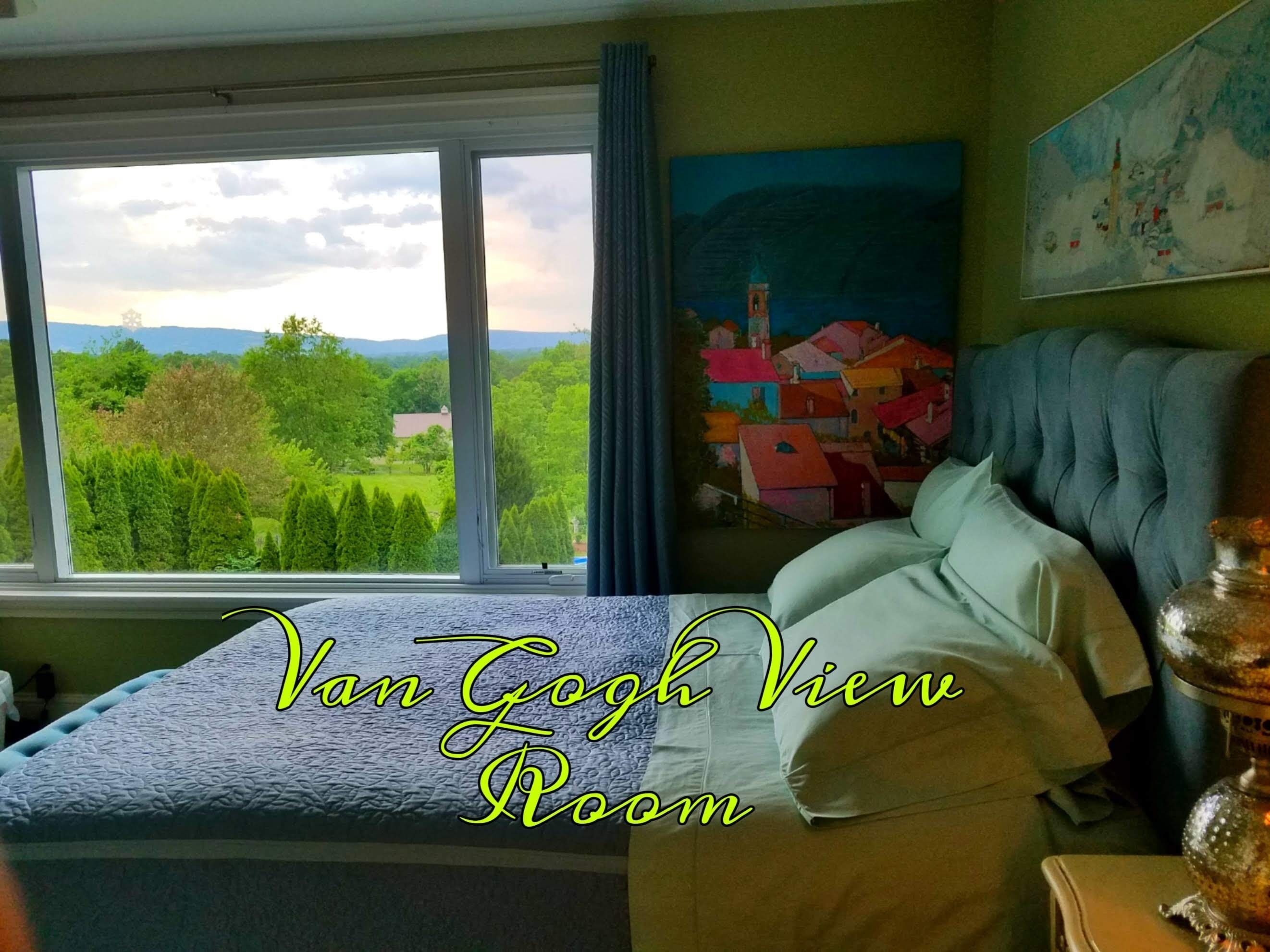 A bedroom with a large window offering a view of a green landscape and a colorful painting on the wall.