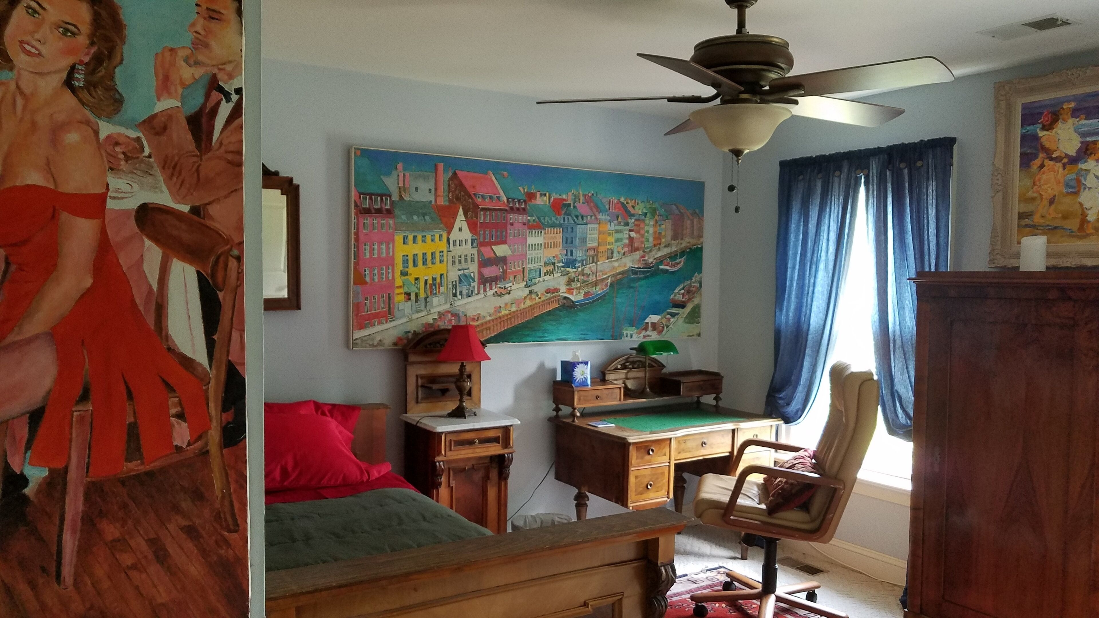 Habitación con cama, escritorio, silla y ventilador de techo. Hay un cuadro en la pared y una ventana con cortinas.