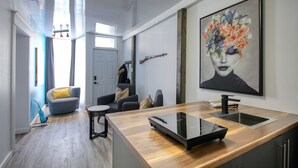 A modern kitchen with a wooden countertop, a built-in oven, and a sink. There is a dining area with a gray sofa and a small round table. A large painting of a floral design is on the wall. - PLACE DES ARTS / ART'S PLACE (Ville de Québec)
