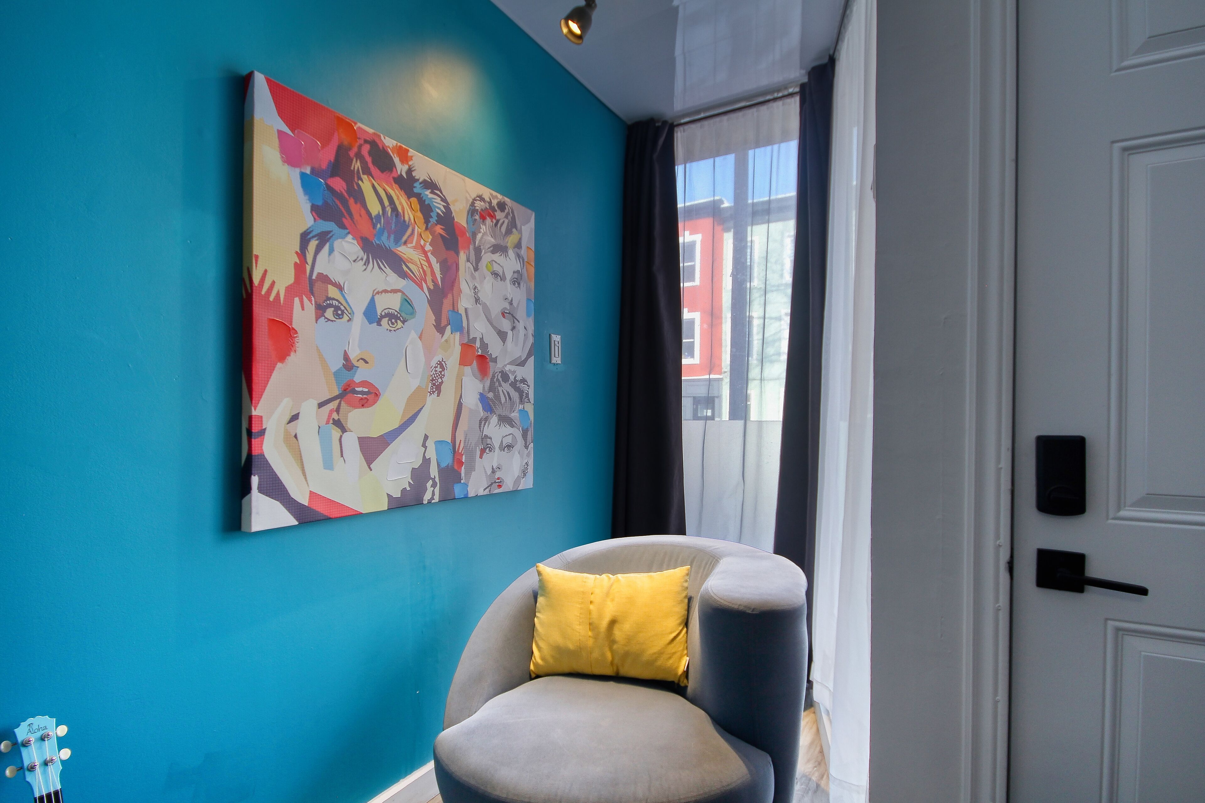 Een kamer met een blauwe wand, een grijze fauteuil met een geel kussen en een kleurrijk abstract schilderij.