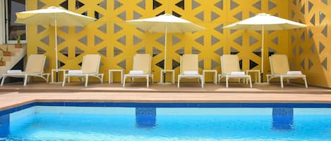 A poolside area with white lounge chairs and umbrellas, a tiled pool, and a yellow patterned wall.