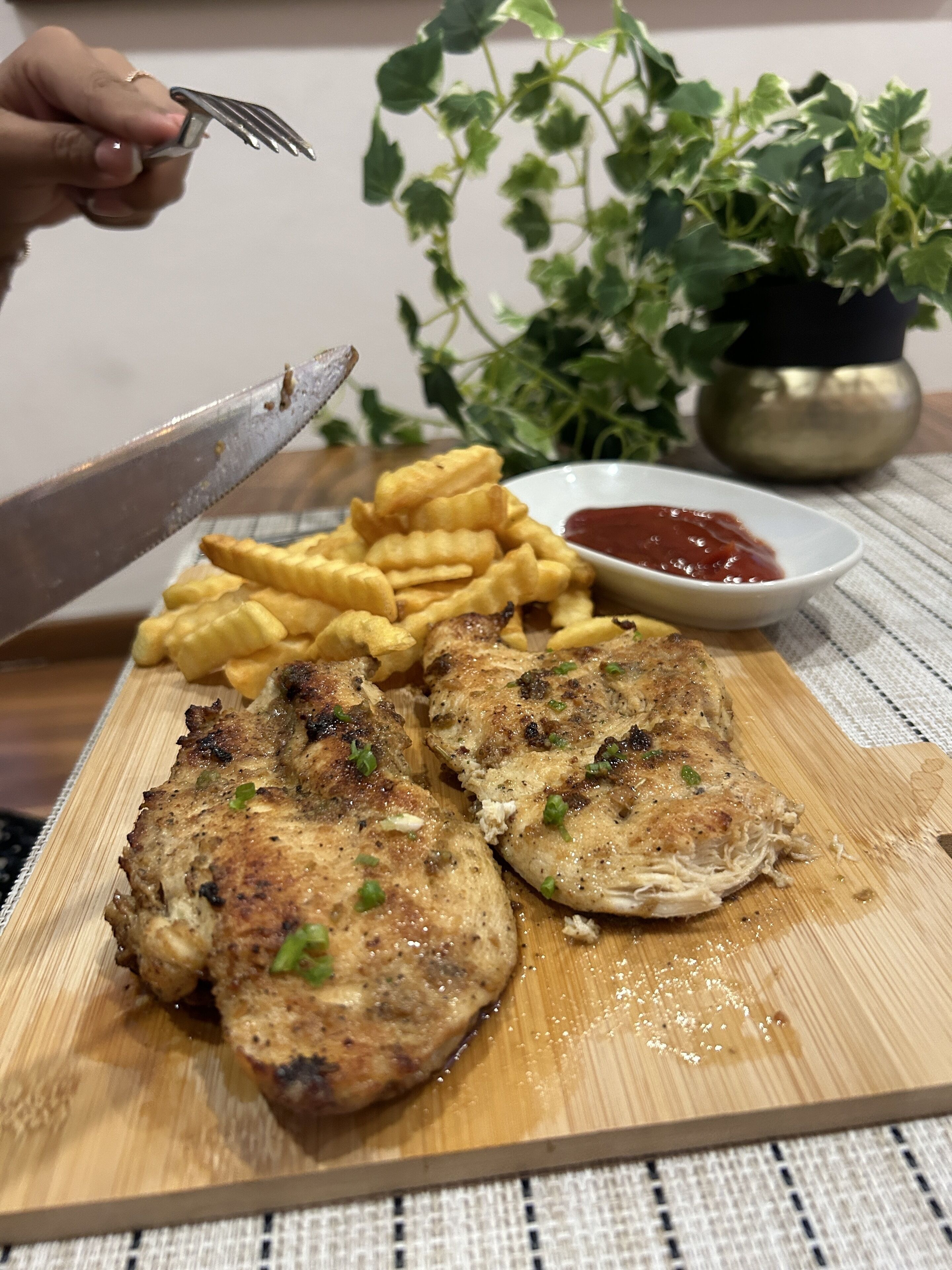 Une planche à découper en bois avec du poulet grillé et des frites, un couteau et un petit bol de ketchup.