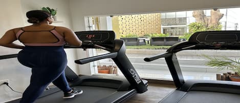 Una cinta de correr en un gimnasio, con una pantalla digital, una ventana que ofrece vistas al exterior y una planta en el alféizar.