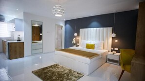 Un dormitorio moderno con una cama grande, mesitas de noche, un pequeño mueble auxiliar, una silla y una cocina visible a través de una puerta abierta.