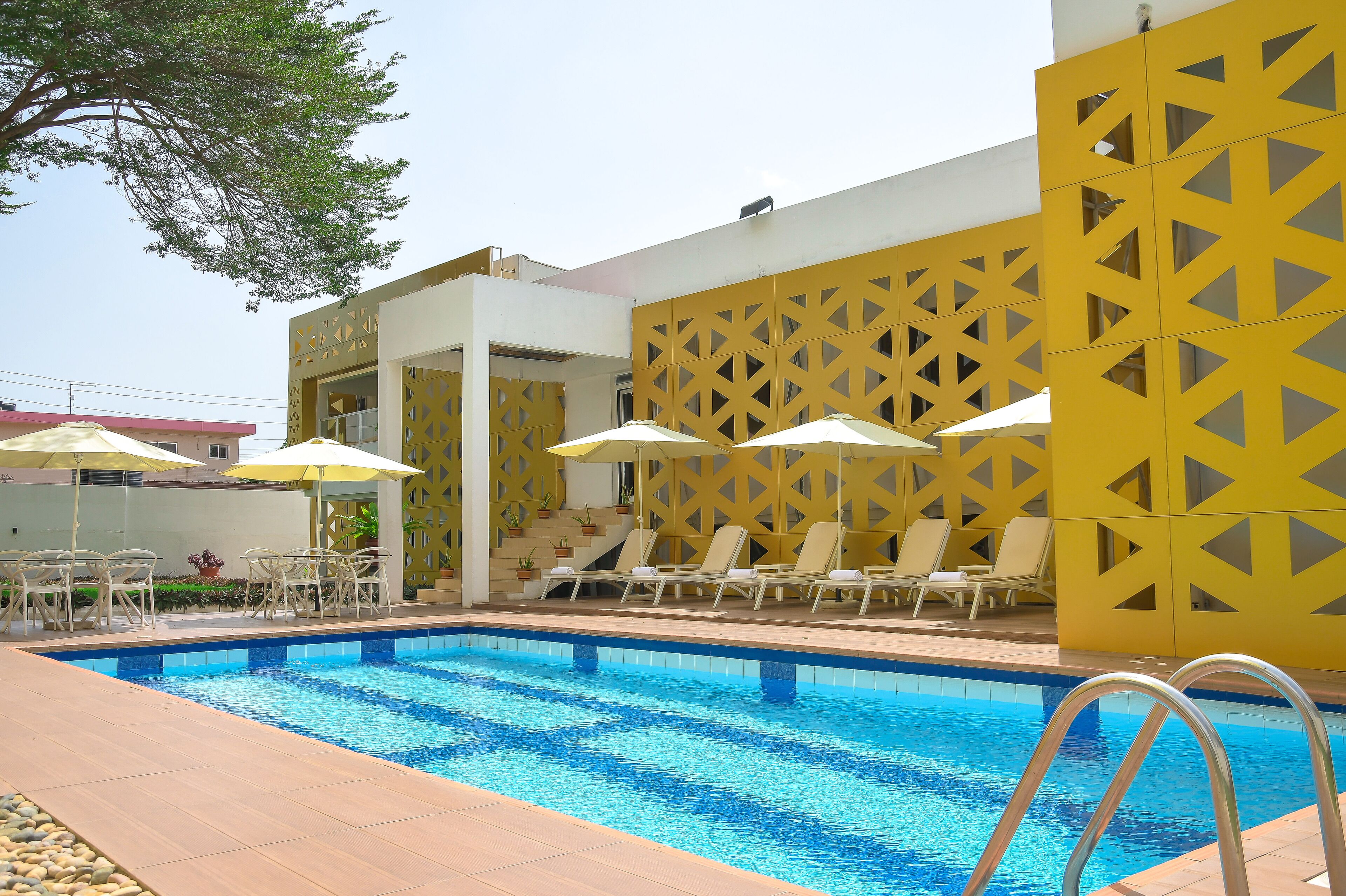 Un espace piscine doté d’une piscine à l’eau d’un bleu limpide, de chaises longues blanches, de parasols jaunes et d’un mur décoratif jaune.