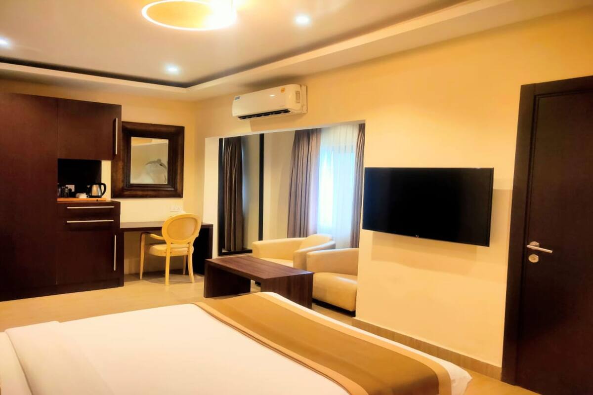 A hotel room with a bed, a desk, a chair, a sofa, a TV, and an air conditioning unit.
