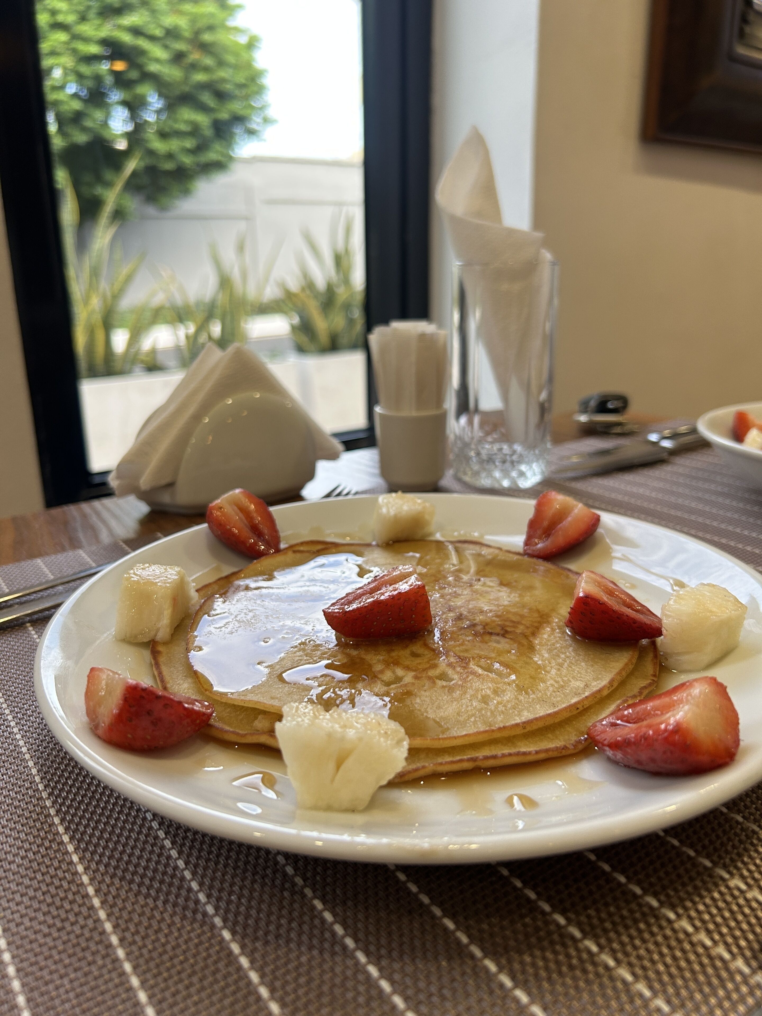 Un plato de desayuno con tortitas, fresas y un vaso de agua sobre una mesa con posavasos.