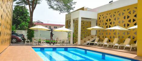 A swimming pool with sun loungers and umbrellas, surrounded by a building with a lattice facade.