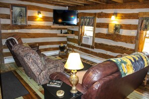 A log cabin interior with a television mounted on the wall, a leather sofa, a patterned armchair, and a lamp on a side table.