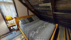 Ein Schlafzimmer mit einem hölzernen Bett, gemusterten Betttextilien, einem Nachttisch mit Lampe und einer schrägen Decke.