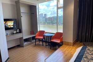 A hotel room with a TV on a shelf, two red chairs around a small table, a large window with a view of buildings and a river, and a bed with a patterned bedspread.