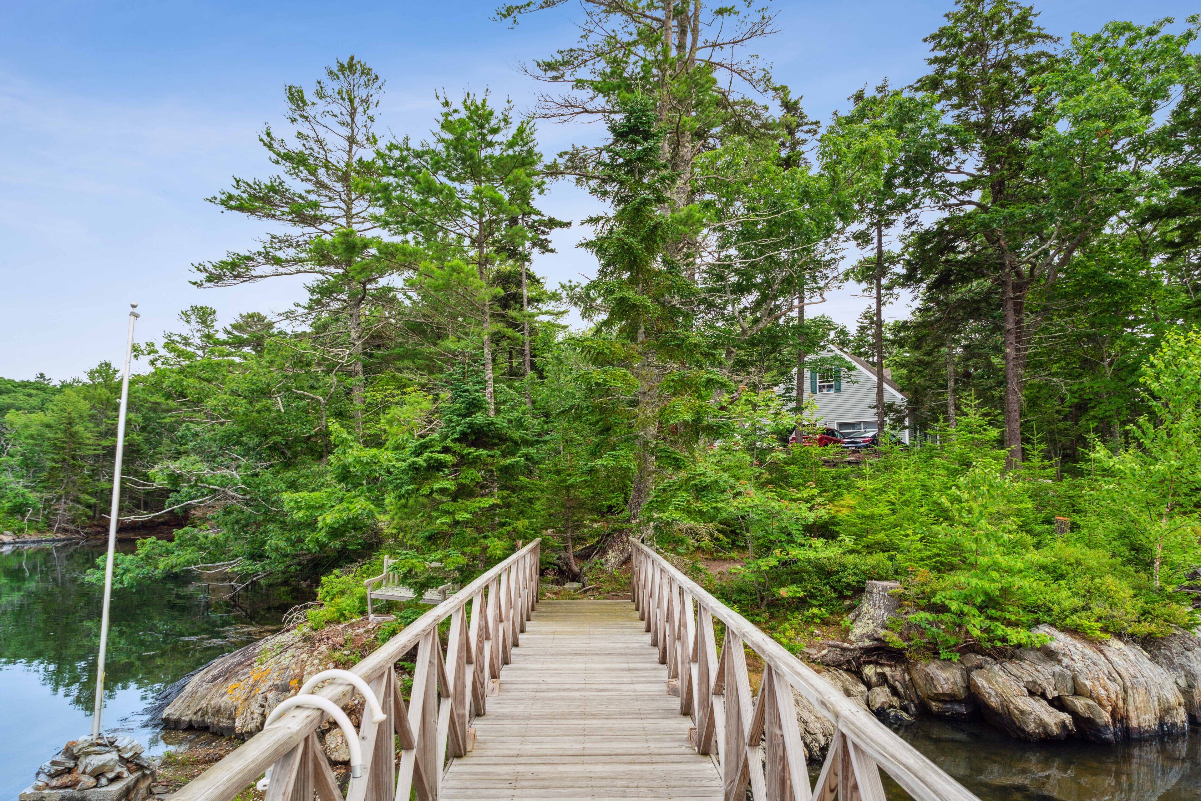 A wooden bridge leading to a house surrounded by dense forest and a body of water.