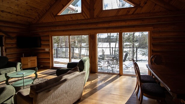 L’intérieur chaleureux d’un chalet avec un plafond en bois, de grandes fenêtres, un coin repas et un espace détente comprenant un canapé et des fauteuils.