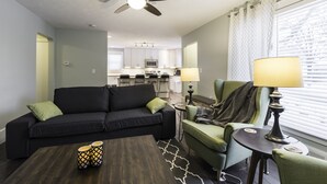 A modern living room with a black sofa, green armchair, and a coffee table. A ceiling fan is visible, and there is a kitchen in the background. - JUST REMODELED top to bottom -  master bed/bath on main floor! (Cleveland Heights)