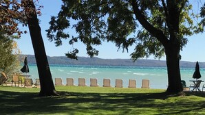 Blick auf einen See mit einer Reihe von Stühlen, die dem Wasser zugewandt sind, umgeben von Bäumen und einer Rasenfläche.