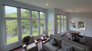Amplia sala de estar con ventanas grandes, un sofá gris, un sillón estampado y un comedor con mesa y sillas.