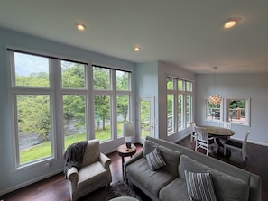 A spacious living room with large windows, a gray sofa, a patterned armchair, and a dining area with a table and chairs.