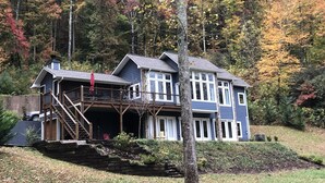 Una casa de dos pisos con balcón y terraza, rodeada por un bosque con follaje otoñal.