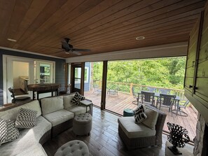 A spacious living area with a large sofa, ottoman, and a dining table with chairs. A ceiling fan is visible, and the space opens to a deck with a view of greenery. - Beautiful, quiet. Right on the river.  New High speed internet! (Hayesville)
