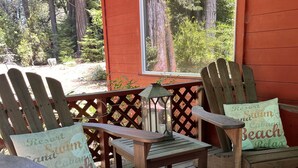 Une terrasse en bois avec des chaises Adirondack et une table, offrant une vue sur une forêt à travers une fenêtre.
