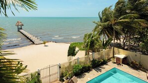 A beach view with a wooden pier leading to a thatched hut, a fenced swimming pool area, and palm trees. - Large 5+ Bedroom Oceanfront w/ Pier and Pool (Placencia)
