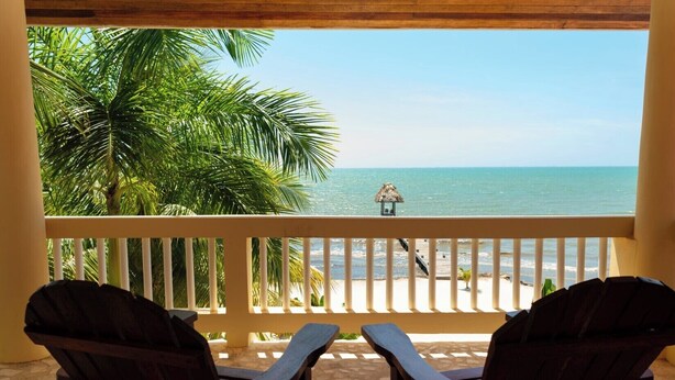 A balcony with two wooden chairs facing a beach with a thatched structure. - Large 5+ Bedroom Oceanfront w/ Pier and Pool (Placencia)