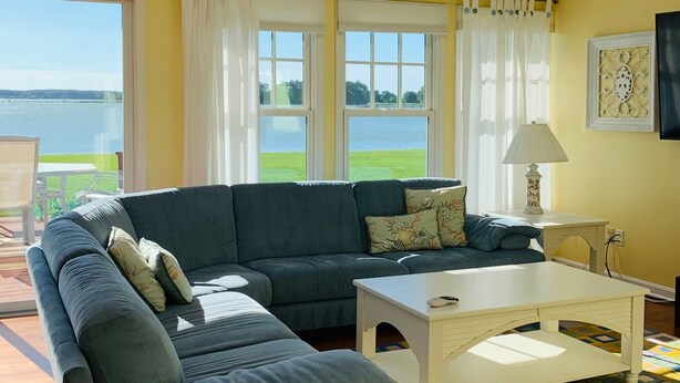 Un salon comprenant un canapé d’angle, un ventilateur de plafond, une petite table et une vue sur un lac, visible à travers de grandes fenêtres.