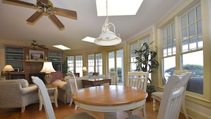 Un espace repas avec une table en bois, des chaises blanches, un ventilateur de plafond et de grandes fenêtres.