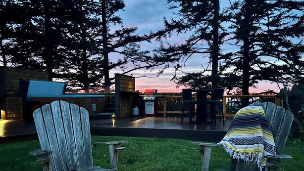 A patio area with a fire pit, two Adirondack chairs, a covered table, and a view of the sunset.