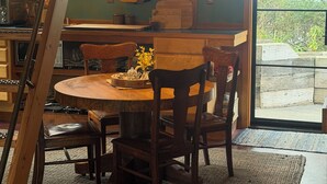 A cozy dining area with a wooden table and chairs, a hanging light fixture, and a view of the outdoors through a large window.