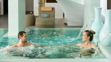 A couple in a modern indoor pool with clear water, a tiled floor, and a large vase on the right.